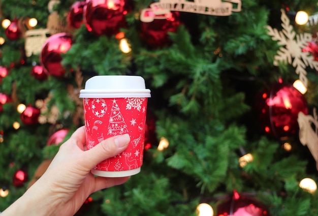 Mão segurando uma xícara de café para viagem com tema de férias com uma árvore de Natal embaçada no fundo