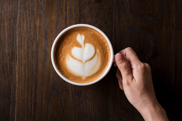 Mão segurando uma xícara de latte art quente na mesa de madeira