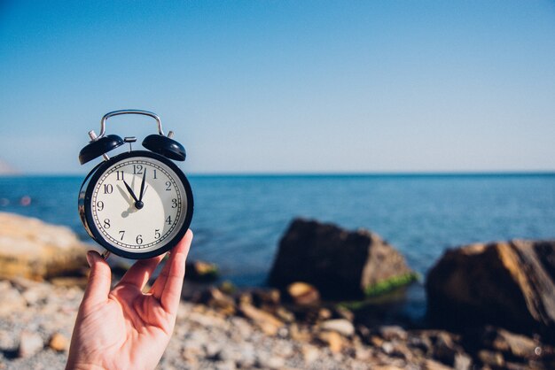 Mão segure o relógio no fundo da praia. Despertador no tempo diferente do fundo do mar e da areia com as férias de verão. Conceito de tempo de férias.