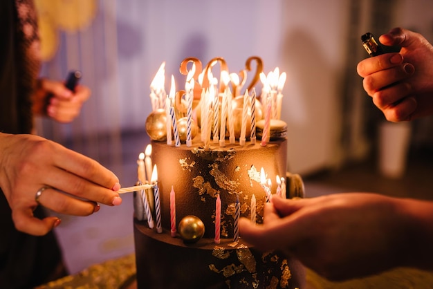 Mãos acendem velas no bolo o bolo de férias é decorado com bolas douradas,  brilhos dourados e macaroons pretos bolo de aniversário masculino 60 anos  fechar