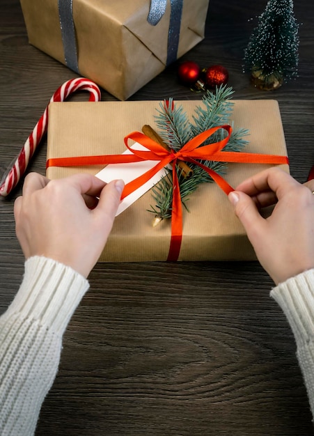 Mãos amarrando uma fita vermelha em uma caixa de presente decorada com um galho de árvore de Natal e um pau de canela em um fundo escuro Copiar espaço