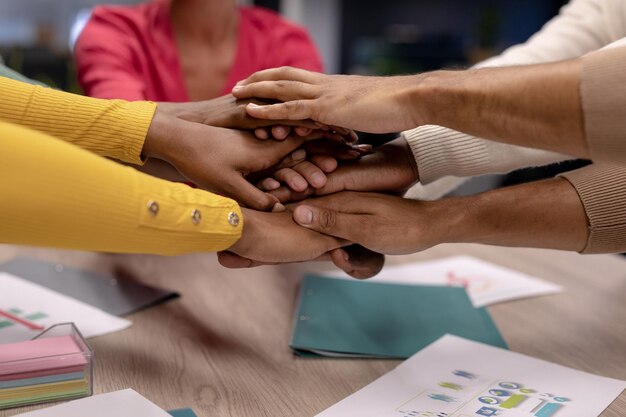 Mãos cortadas de colegas birraciais do sexo feminino e masculino empilhando as mãos sobre a mesa de conferência no escritório