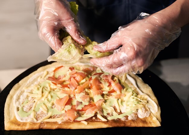 Mãos cortadas do chef rasgando alface na base assada redonda Pedaços picados de ingredientes para cozinhar pizza Vista de perto