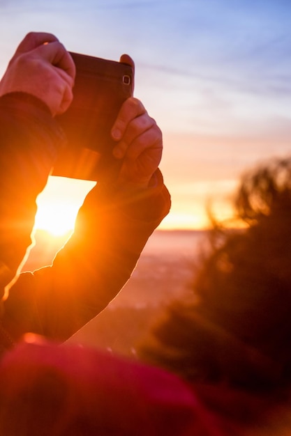 Foto mãos cortadas fotografando de telefone móvel contra o céu durante o pôr do sol