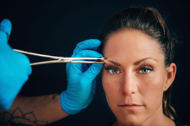 Foto mãos cortadas perfurando sobrancelha de mulher contra fundo preto