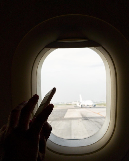 Foto mãos cortadas usando telefone pela janela de um avião.