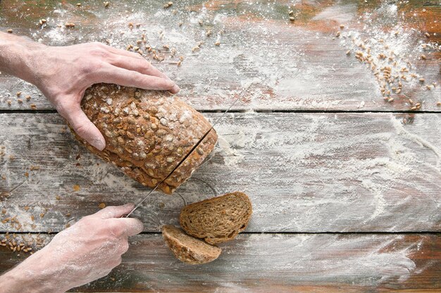 Mãos cortando pão com faca na mesa de madeira rústica polvilhada com farinha.