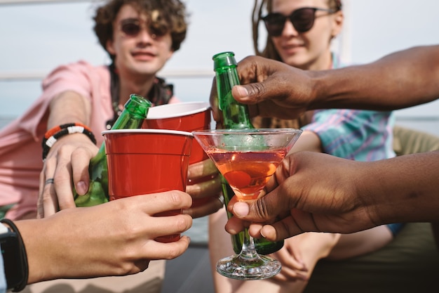 Mãos de amigos brindando com garrafas de cerveja, copos de plástico e copos de martini na festa