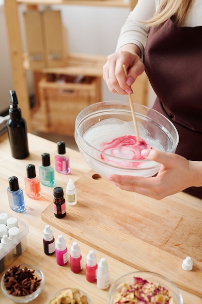 Mãos de artesã misturando massa de sabonete líquido com cor carmesim em vidro com uma vara de madeira sobre a mesa