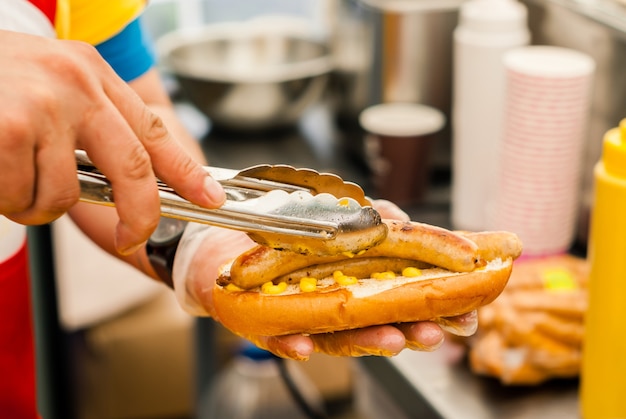 Foto mãos de chef de rua com pinça colocando duas salsichas em um pão de perto