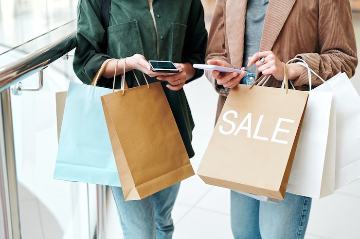 Foto mãos de dois jovens compradores contemporâneos com smartphones e sacos de papel navegando por produtos on-line e comparando preços