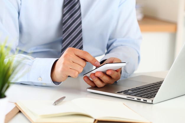 Mãos de empresário digitando algo no telefone inteligente, sentado em seu escritório. Vista de perto