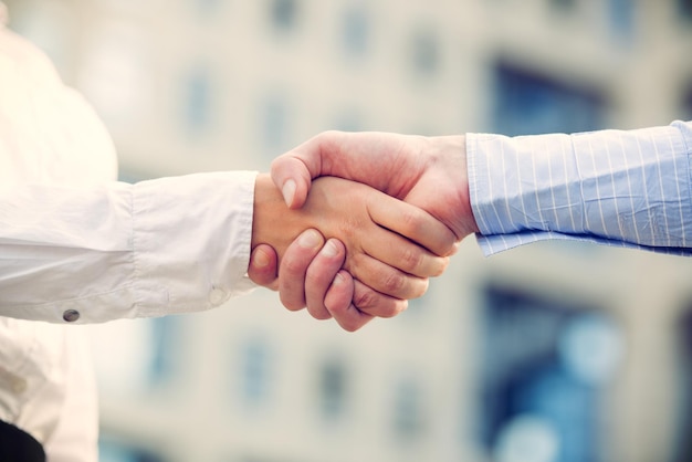 Mãos de empresário e empresária apertando as mãos, enquanto no fundo aparece o prédio de escritórios.