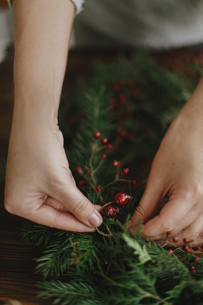 Mãos de florista segurando bagas e fazendo guirlanda de natal rústica na mesa de madeira