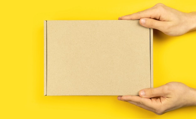 Mãos de homem entregador com maquete de caixa de papelão, vista superior. Fundo amarelo da mesa. Conceito de envio e entrega, cópia da foto do espaço