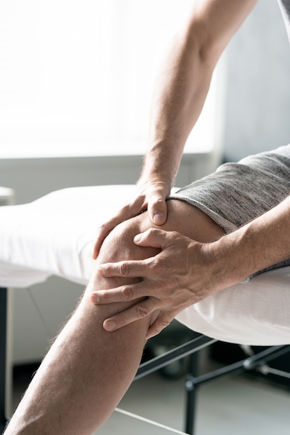 Mãos de homem maduro massageando seu joelho direito enquanto está sentado na mesa para procedimentos fisioterapêuticos em clínicas ou consultórios médicos