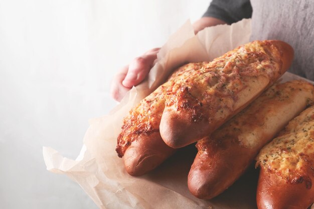 Mãos de homem segurando sanduíche quente no pão baguete com presunto bacon legumes e queijo em pergaminho Macho de avental preto no fundo da cozinha em casa com padaria Mock up