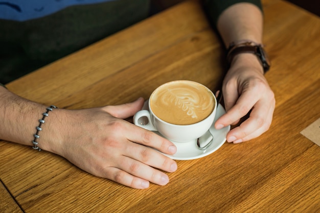 Mãos de homem segurando uma xícara de cappuccino, café, mesa de madeira