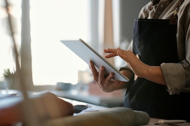 Mãos de jovem alfaiate ou designer de roupas usando tablet na oficina