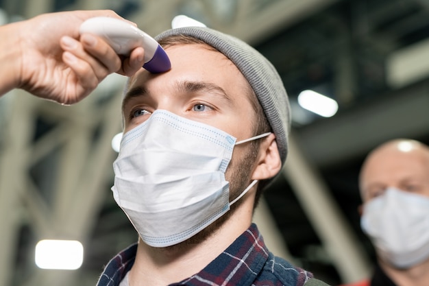 Mãos de jovem trabalhador da área médica com termômetro sem contato medindo a temperatura corporal de engenheiros com máscaras protetoras antes do trabalho