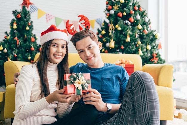 Mãos de jovens asiáticas seguram um presente de Natal em uma caixa vermelha com fita com o namorado em uma sala de estar com uma árvore de Natal decorada ao fundo