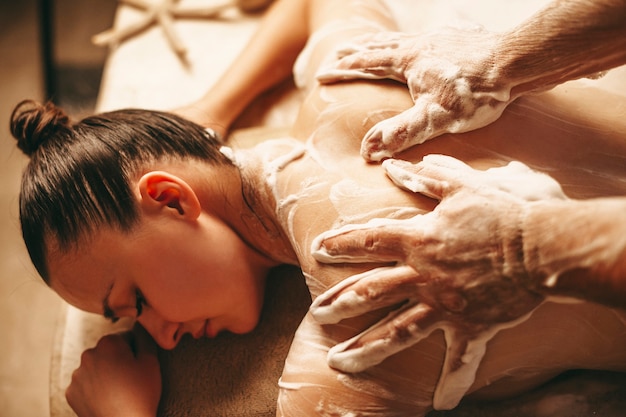 Mãos de massagista fazendo uma massagem de espuma nas costas de uma mulher em um centro de spa de bem-estar.