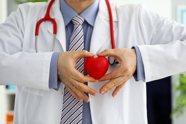 Mãos de médico de medicina masculina segurando e cobrindo coração de brinquedo vermelho