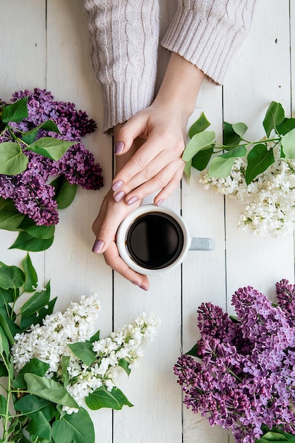 Mãos de menina com uma xícara de café na mesa com flores lilás postura plana