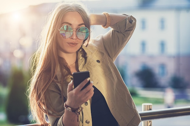 Mãos de mulher bonita e elegante segurando um telefone fundo do pôr do sol dia ensolarado bom tempo óculos de sol acessórios legais