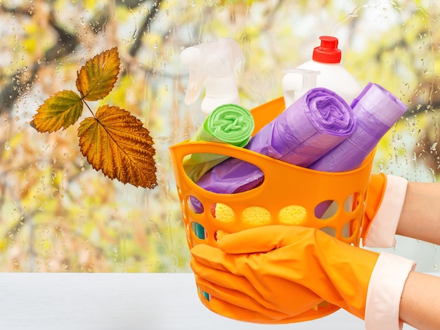 Mãos de mulher com luvas de proteção de borracha segurando uma cesta de laranja com sacos de lixo, garrafas de vidro e limpador de azulejos em frente à janela com gotas de água e folhas de outono. Conceito de limpeza.