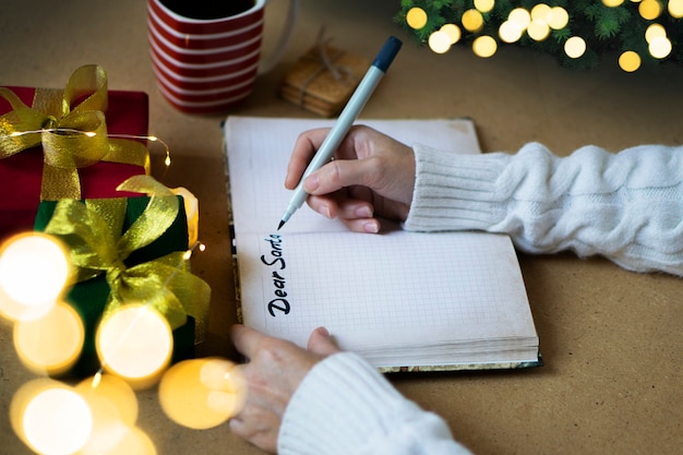 Foto mãos de mulher com suéter branco escrevendo carta para o papai noel no caderno, foco seletivo, luzes de bokeh, foto atmosférica. conceito de compras e presentes de natal.
