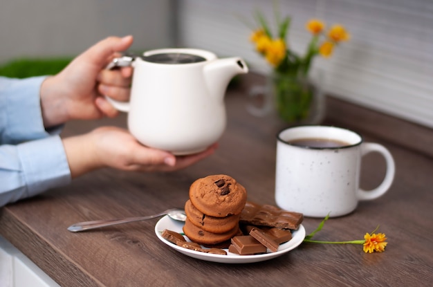 Mãos de mulher com xícara de chá de chaleira branca e biscoito de chocolate