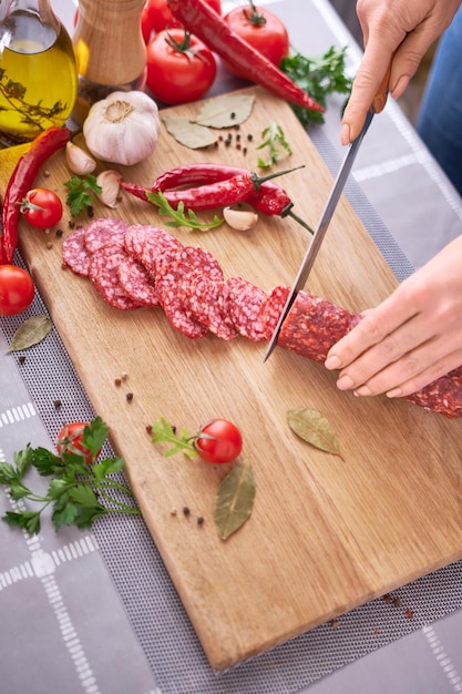 Mãos de mulher cortando uma salsicha de salame na tábua de madeira na cozinha doméstica