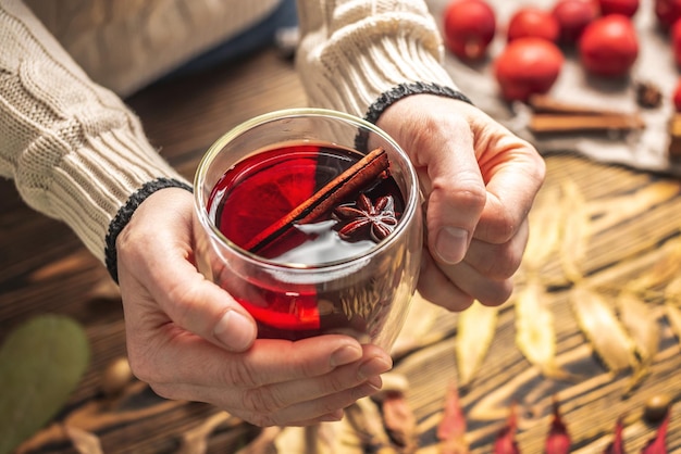 Mãos de mulher de suéter estão segurando um copo de vidro com vinho quente em um fundo de madeira com folhas de outono Conceito de uma bebida tradicional de aquecimento