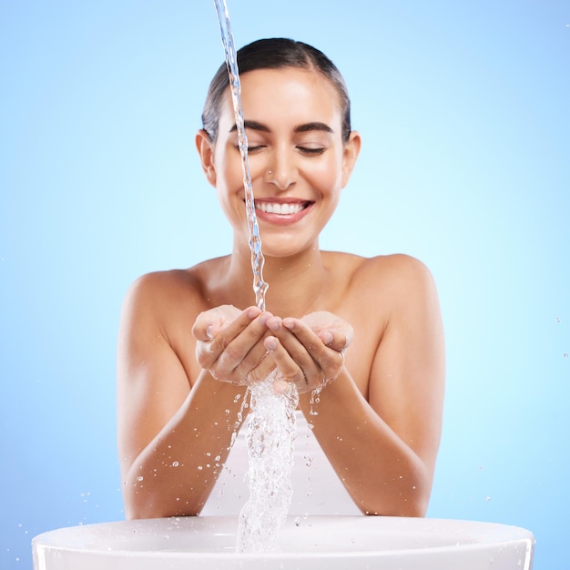 Foto mãos de mulher e respingos de água com sorriso para lavagem de higiene da pele ou tratamento facial na pia contra o fundo azul do estúdio fêmea feliz sorrindo em beleza natural limpar minerais ou autocuidado