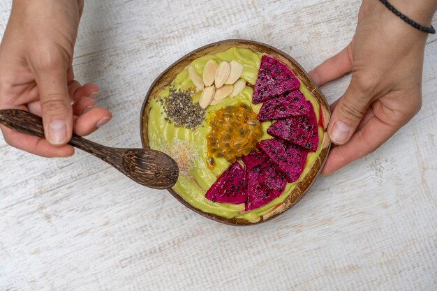Mãos de mulher e suco de abacate verde em uma tigela de coco com fruta do dragão, maracujá, flocos de amêndoa, lascas de coco e sementes de chia no café da manhã. Conceito de alimentação saudável, superalimento. Bali, Indonésia