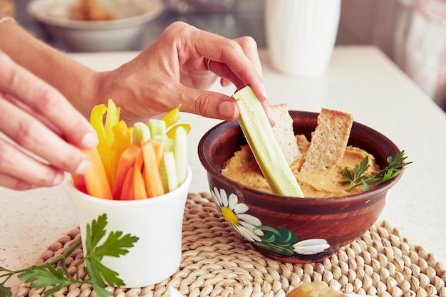 Mãos de mulher mergulham pepino fresco em homus caseiro com biscoitos salsa e legumes frescos na mesa Lanches rústicos vegetarianos saudáveis fecham