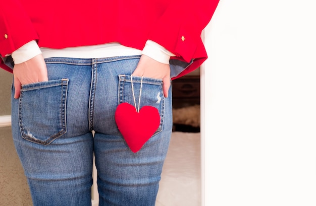 Mãos de mulher nos bolsos de trás da calça jeans com grande coração vermelho do dia dos namorados perto da porta branca.