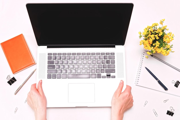 Mãos de mulher segurando laptop. Espaço de trabalho com diário e flores sobre fundo branco. Postura plana, vista superior