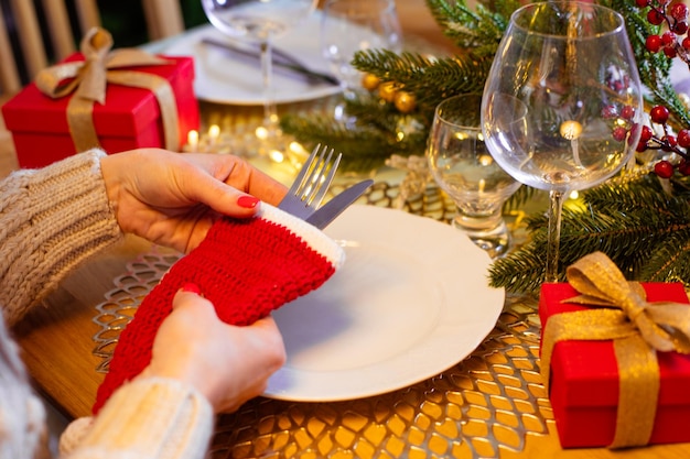 Mãos de mulher segurando um pequeno chapéu de Papai Noel de crochê com talheres Usando decoração artesanal para tornar as férias especiais Cenário de mesa festiva com detalhes vermelhos e dourados