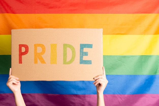 Mãos de mulher segurando uma placa de papelão com a palavra orgulho escrita com as letras coloridas e a bandeira do arco-íris atrás