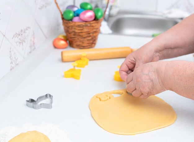 Mãos de mulher sênior cortadas de biscoitos de massa para o dia da páscoa feliz conceito de páscoa processo de fazer biscoitos cozinhar em casa conceito tradição comida caseira