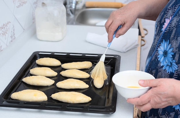 Mãos de mulher sênior escovando tortas cruas com ovo antes de assar em uma assadeira Foco seletivo Processo de fazer tortas com recheio de maçã Conceito de cozinhar em casa Tradição comida caseira