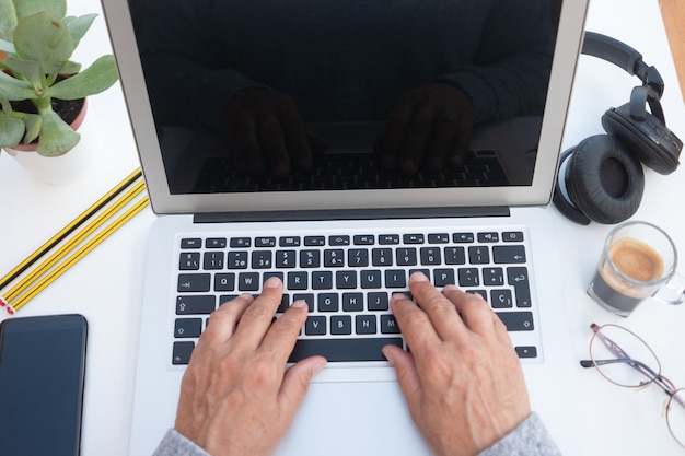 Mãos de mulher sênior, trabalhando no laptop. Mesa branca. Um café para fazer uma pausa. Escritório alternativo ao ar livre. Celular e fones de ouvido perto dela