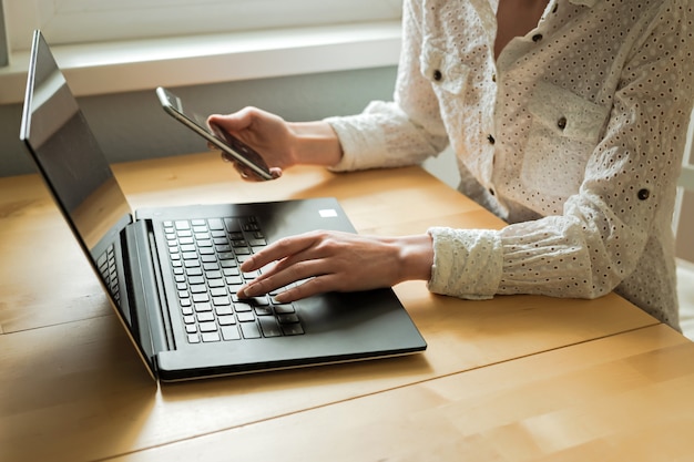 Mãos de mulher usando laptop e smartphone na mesa no interior de casa