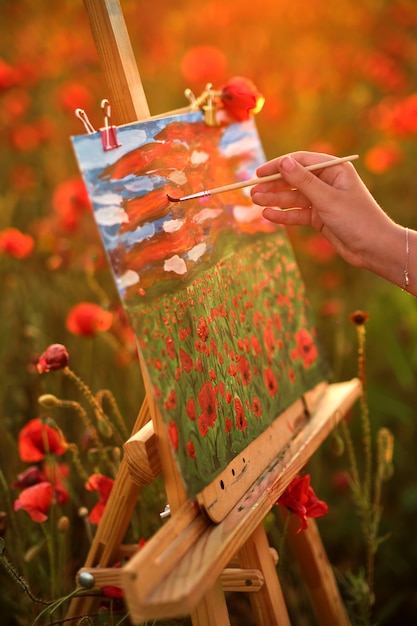 Mãos de um artista com pincel fecham em um campo de papoulas vermelhas ao pôr do sol