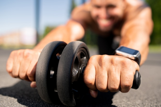 Mãos de um forte atleta mascular segurando as alças do aparador enquanto se exercita em um campo esportivo ao ar livre