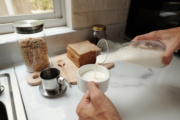 Mãos de um homem derramando leite na xícara de café ao preparar o café da manhã