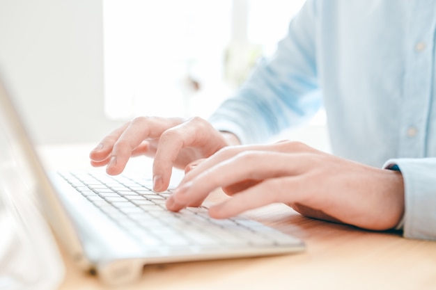 Mãos de um jovem analista ou designer digitando em um pequeno teclado branco enquanto está sentado à mesa na frente do computador