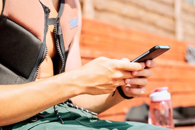 Mãos de um jovem ativo em uma jaqueta esportiva de segurança e shorts enviando mensagens ou rolando no smartphone enquanto descansam entre os treinamentos no resort de verão
